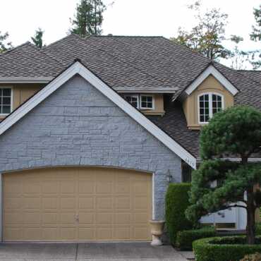 A suburban house with a stone facade and brown roof features a two-car garage with a yellowish door. A manicured shrub is in the foreground, and trees are visible in the background.