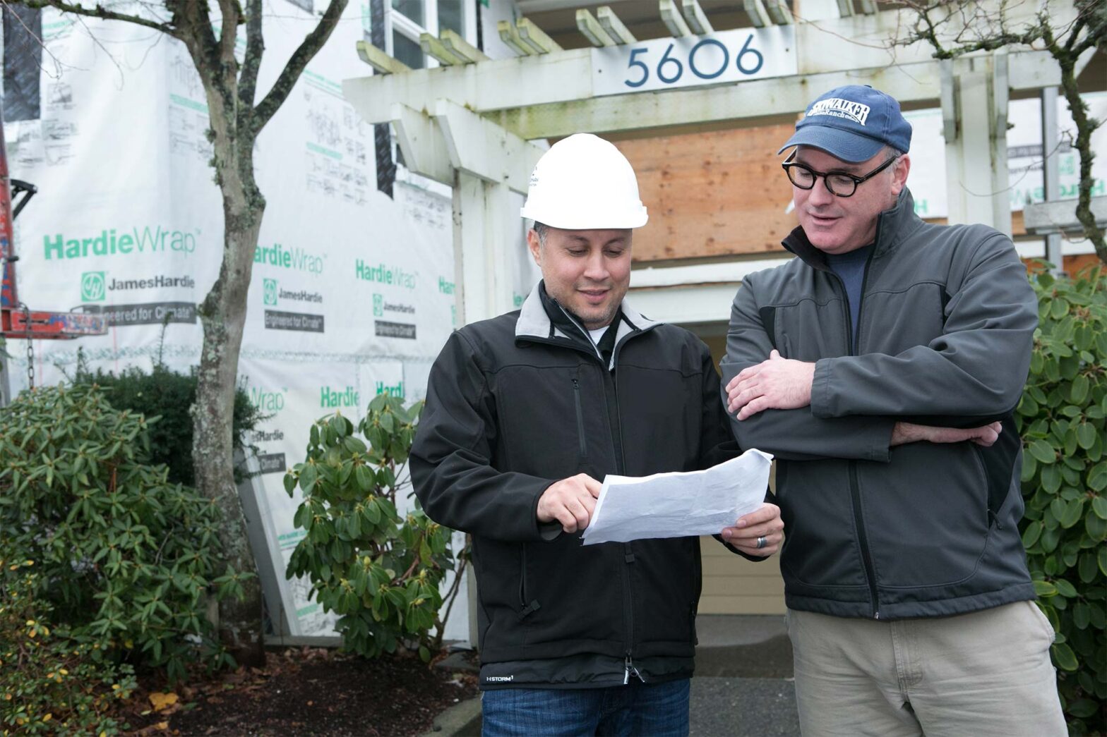 Two men outside a house under construction. One wears a white hard hat and holds a document, while the other wears glasses and a cap. They appear to be discussing the plans. The house is partially wrapped in construction material.