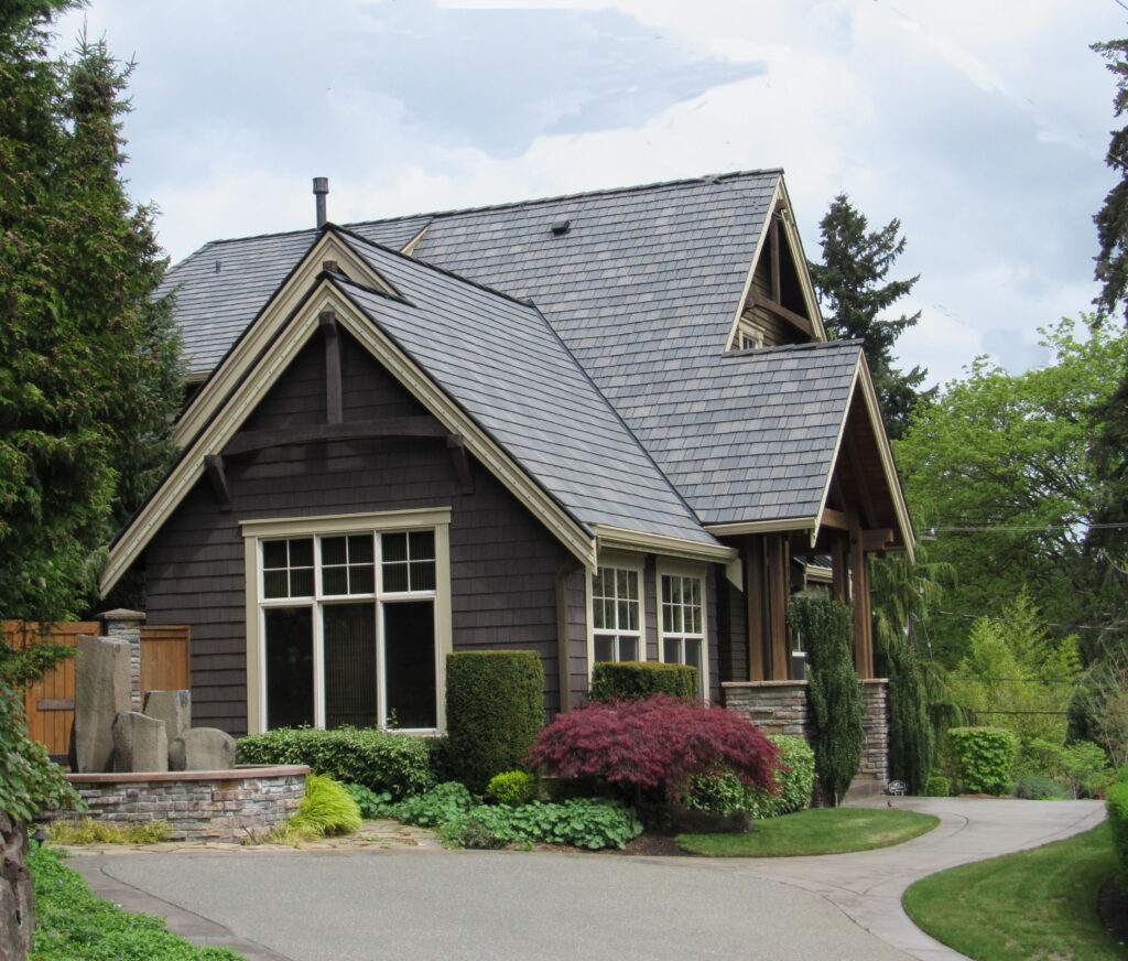 A modern cottage style house with dark wood siding, large windows, and a steep roof, surrounded by lush greenery, shrubs, and a curved driveway under a cloudy sky.