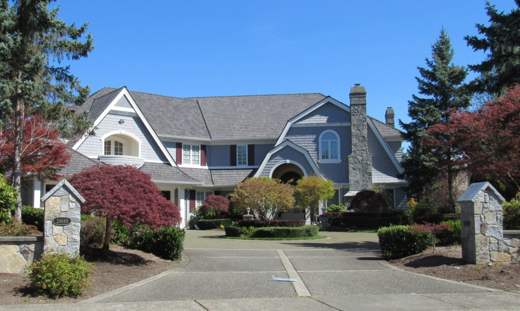 A large, two story suburban house with gray siding, white trim, and a stone chimney. It is surrounded by manicured landscaping, bushes, and trees, with a wide driveway leading to the entrance.