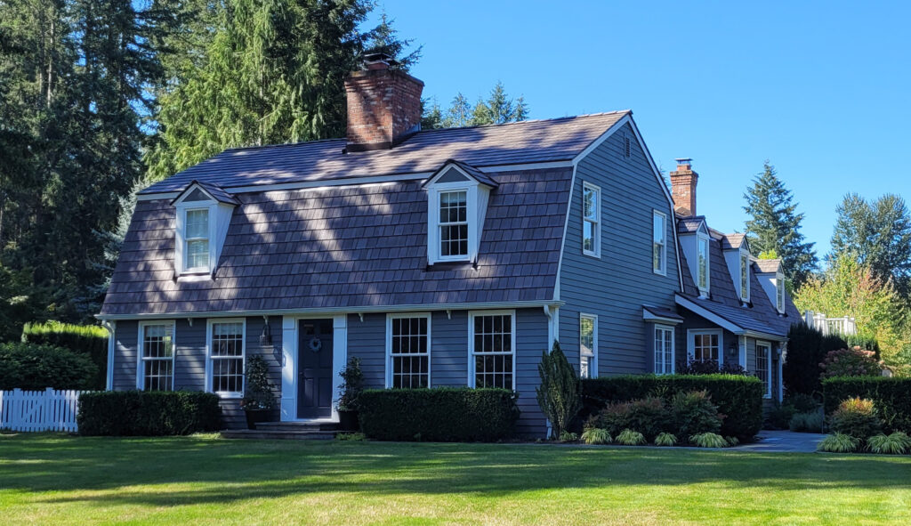 A two story blue house with a steep, shingled roof, white trim, dormer windows, and brick chimneys sits on a well manicured lawn with shrubs and trees around it on a sunny day.