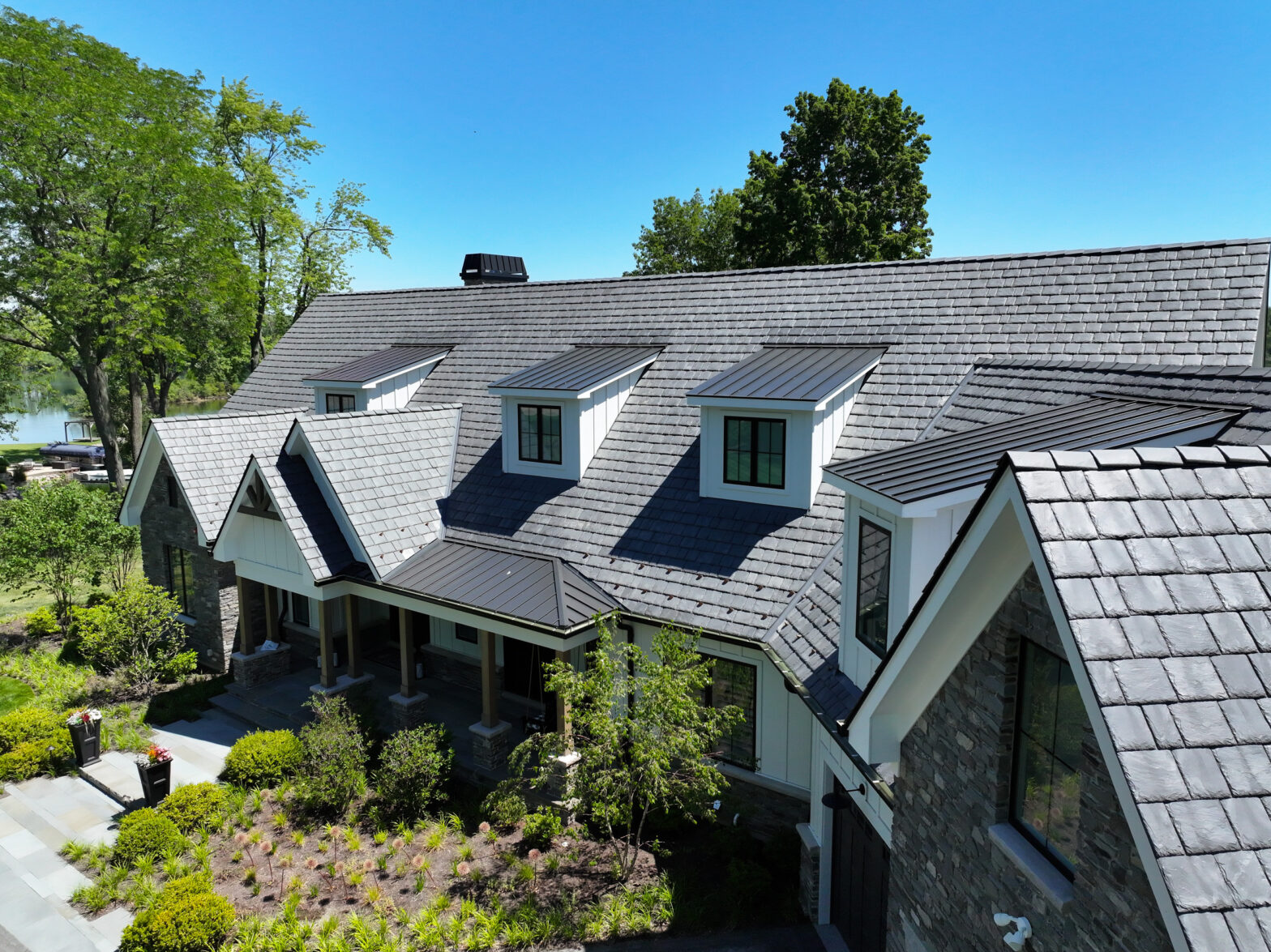 A large house with gray shingle roofing, white dormer windows, and stone exterior, surrounded by landscaped greenery, under a clear blue sky.