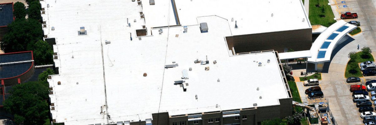 Aerial view of a large white-roofed building surrounded by parked cars and green lawns. The structure has an irregular shape, and there are trees and other buildings nearby. Pathways and driveways are visible around the premises.
