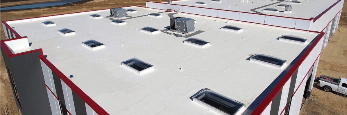 Aerial view of a large industrial building with a white flat roof and red trim. Several skylights and HVAC units are visible. The building is surrounded by flat, open fields under a clear blue sky.